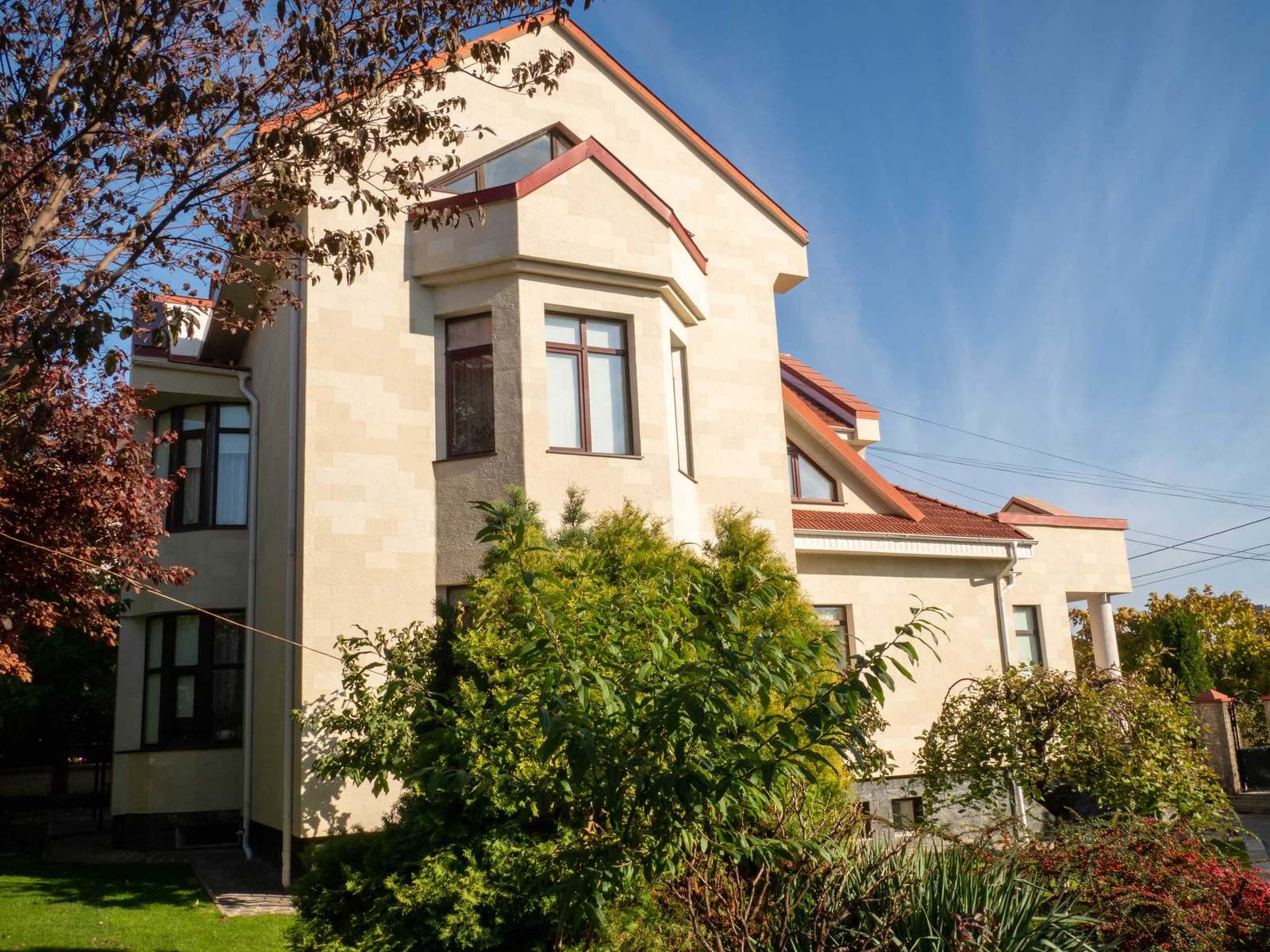 La maison vue du jardin, avec bow-window, haut pignon et façade en pierre flexible beige parmi les arbustes